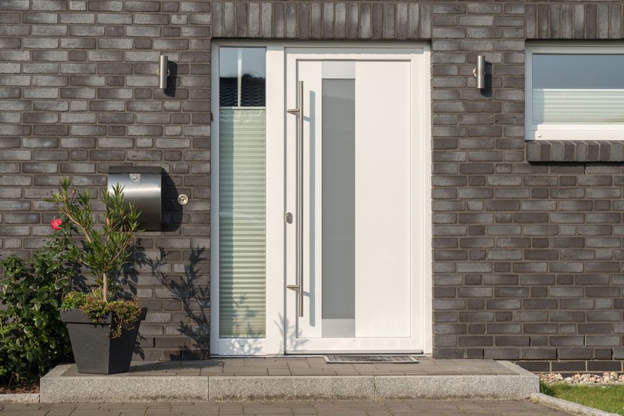 Vordereingang eines modernen Hauses mit weißer Tür, grauer Backsteinmauer und Topfpflanze neben der Treppe