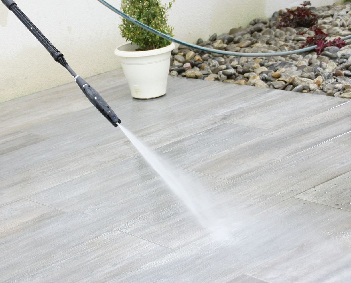 Un nettoyeur haute pression nettoie une terrasse carrelée grise en pulvérisant de l'eau.
