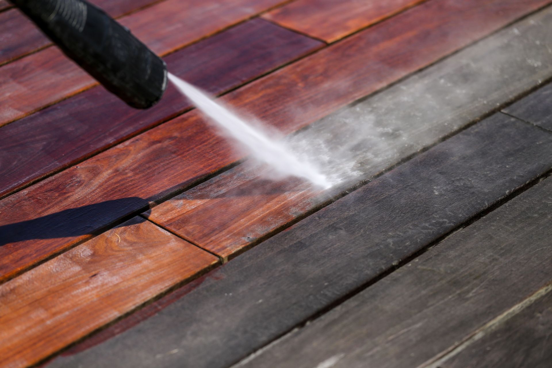 Nettoyeur haute pression pulvérisant un jet d'eau sur une terrasse en bois, enlevant la saleté et révélant des variations de couleur.
