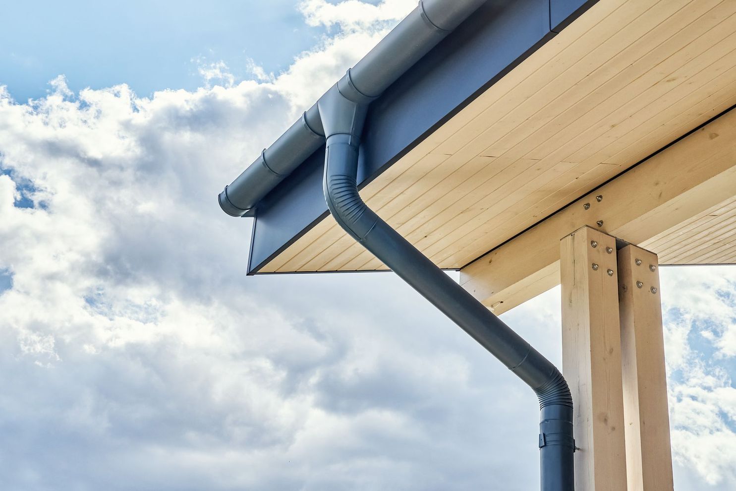 Gouttière grise à l'angle d'un bâtiment en bois clair, sous un ciel bleu nuageux.