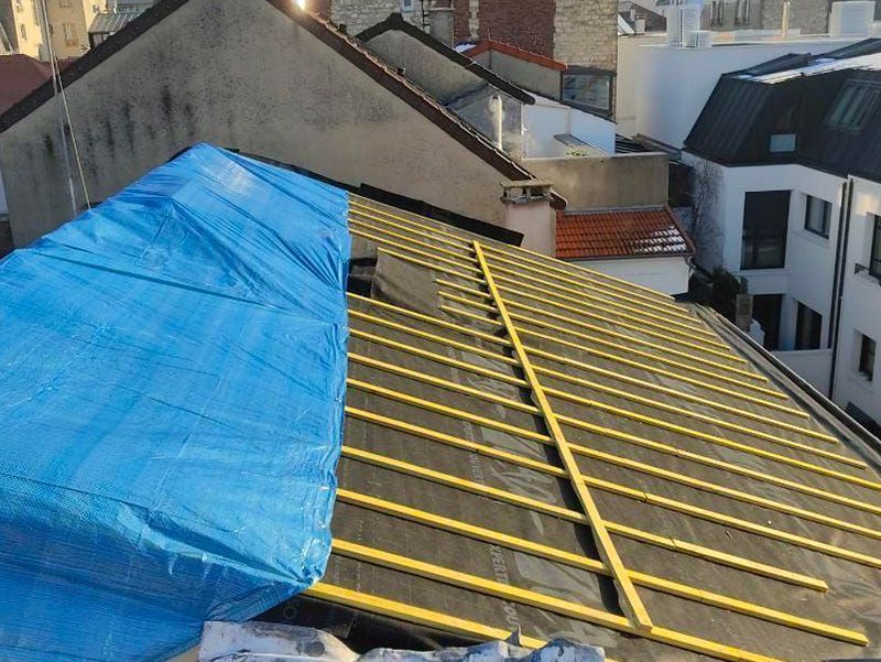 Toiture en travaux ; une bâche bleue en recouvre une partie. On aperçoit des poutres en bois et une sous-couche sombre. Des bâtiments se dressent à l’arrière-plan.