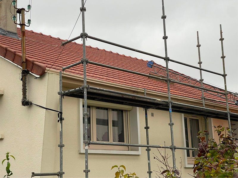 Des échafaudages sont installés près d'une maison au toit de tuiles rouges, aux murs et fenêtres couleur crème.