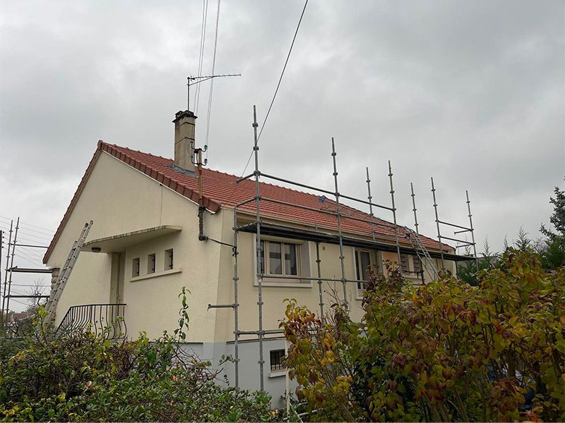 Extérieur de la maison avec échafaudages pour travaux de toiture, ciel couvert. Bardage beige, toit de tuiles rouges.