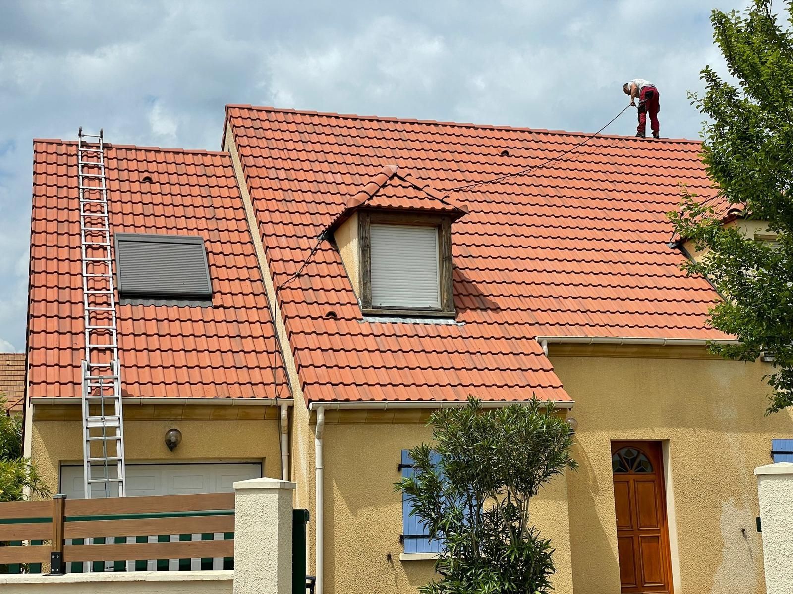 Un homme travaille sur un toit de tuiles rouges. Une échelle est appuyée contre le mur. Maison jaune, ciel nuageux.