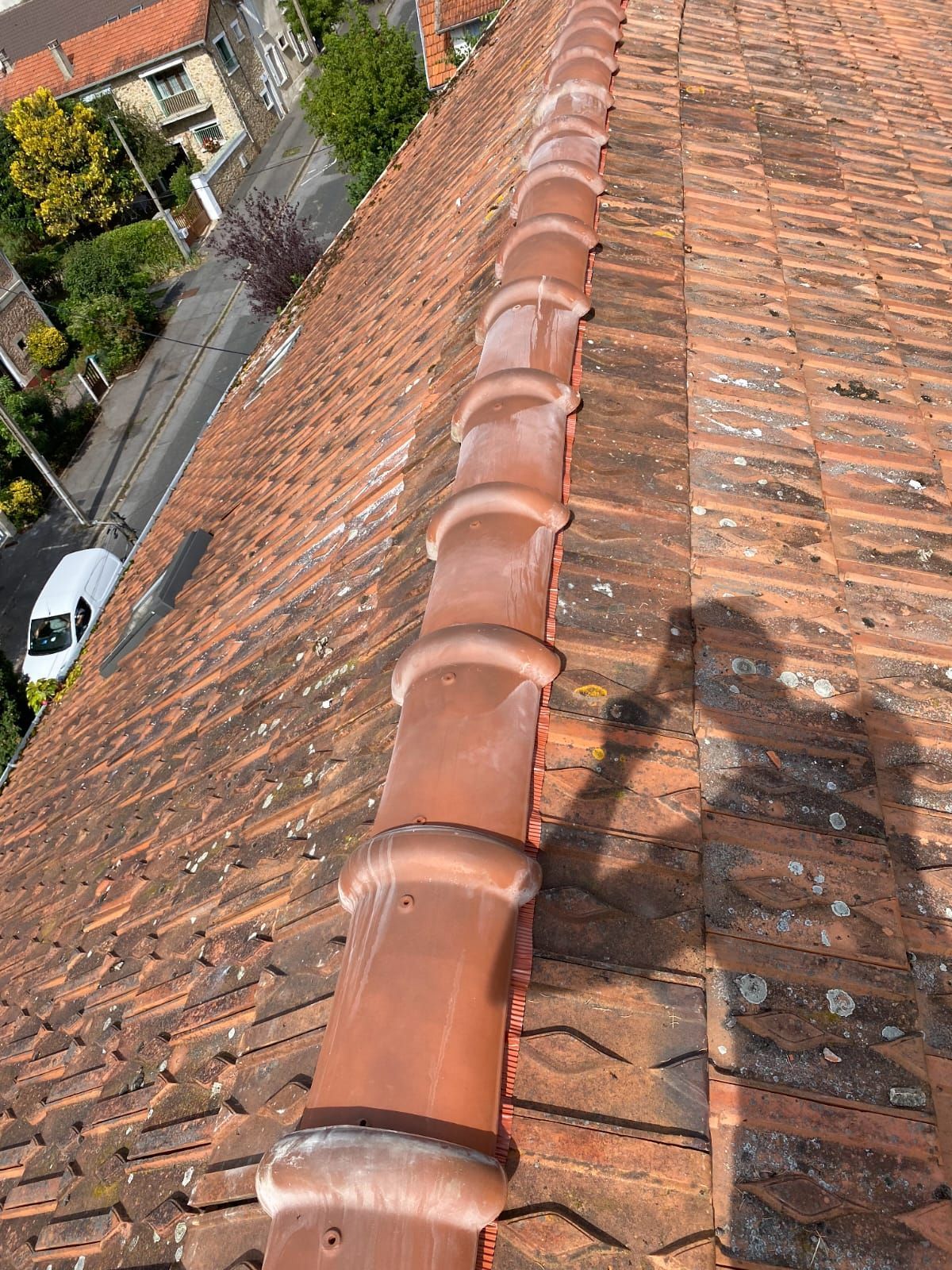 Gros plan sur un toit de tuiles rouges avec une longue gouttière couleur cuivre. La vue inclut une rue et des bâtiments en contrebas.