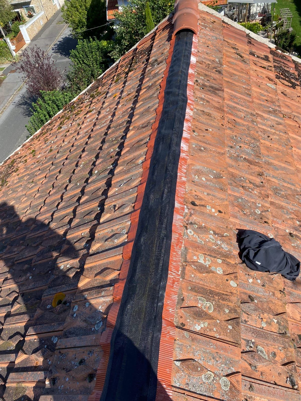 Toit de tuiles rouges avec un grillage noir le long du faîtage, probablement pour empêcher les oiseaux d'y pénétrer, avec une ville visible en arrière-plan.