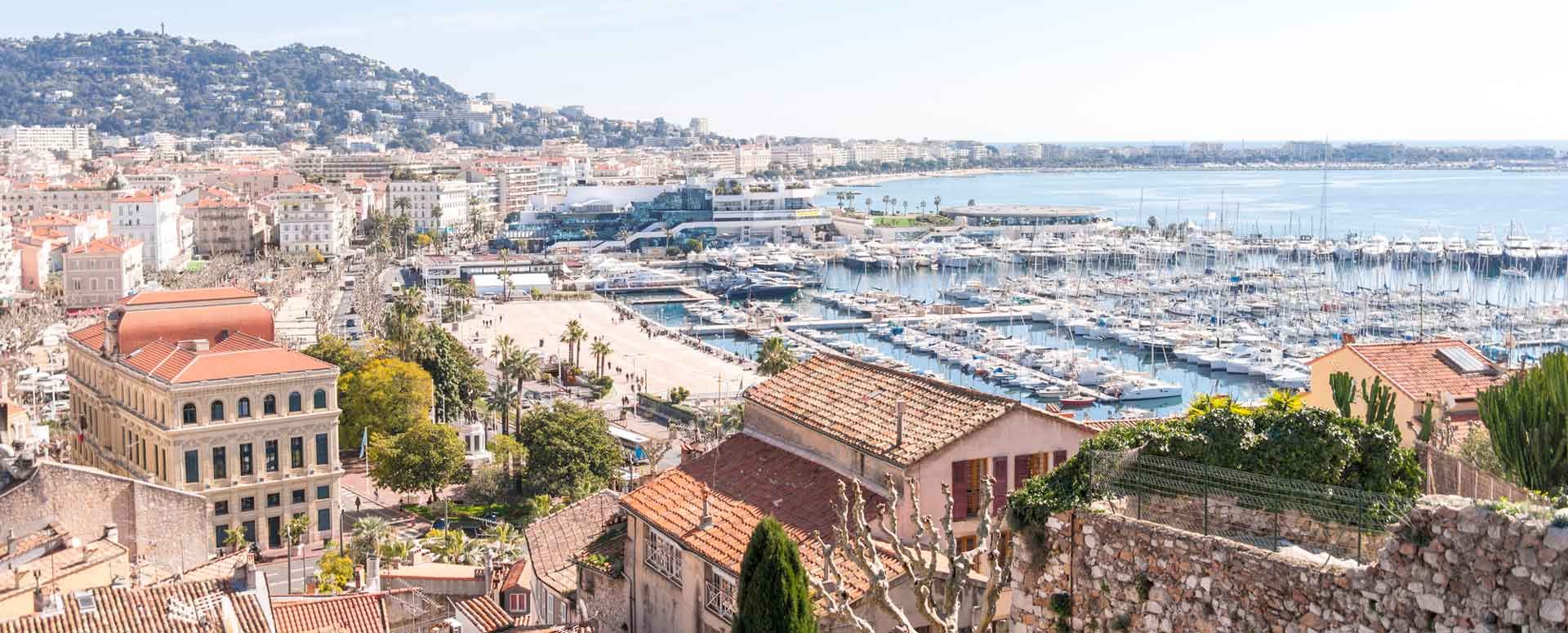 Vue panoramique de Cannes, en France. Bâtiments, port avec de nombreux bateaux, mer bleue et colline en arrière-plan.