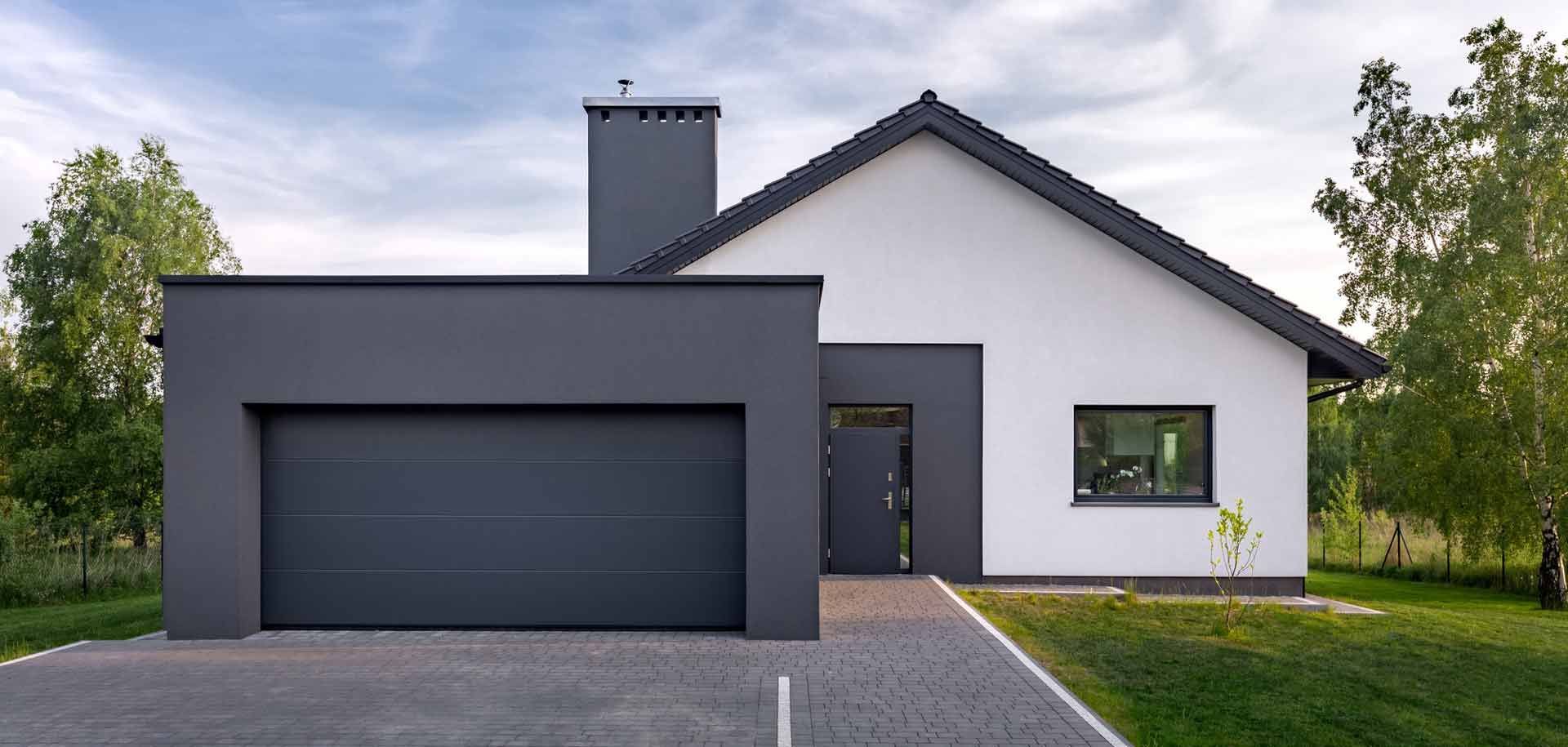 Maison moderne avec garage gris et façade blanche, allée en briques, pelouse verte et arbres.