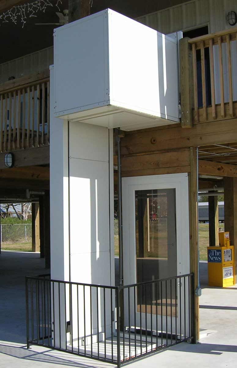 Ascenseur avec cabine blanche et rambarde de sécurité, à côté d'un bâtiment en bois, sur une dalle en béton.