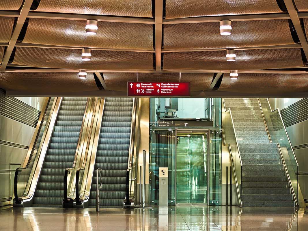 Escaliers mécaniques, ascenseur et escaliers dans un immeuble moderne. Plafond marron avec éclairage. Enseigne rouge au-dessus.