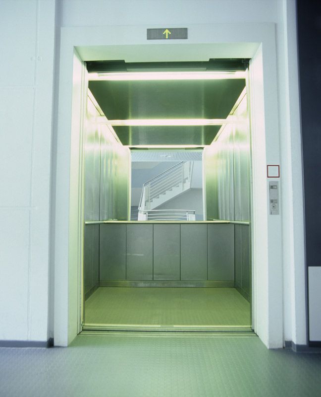 Ascenseur vide, portes ouvertes, donnant sur un escalier d'un immeuble. L'éclairage est assuré par des tubes fluorescents.