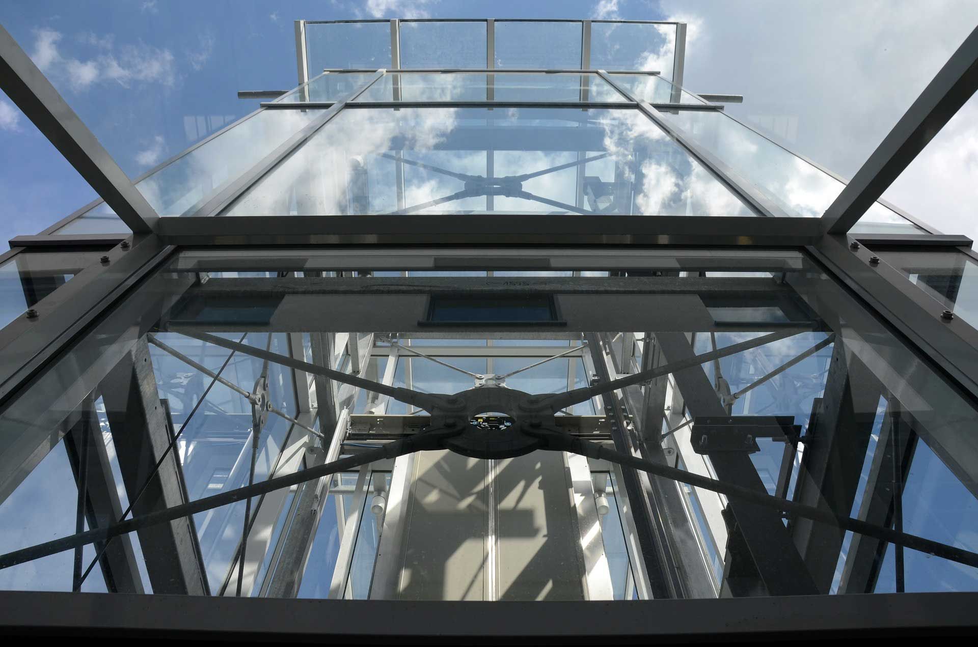 En levant les yeux à travers une structure de verre et de métal, on aperçoit une tour avec des poutres de soutien et le ciel.