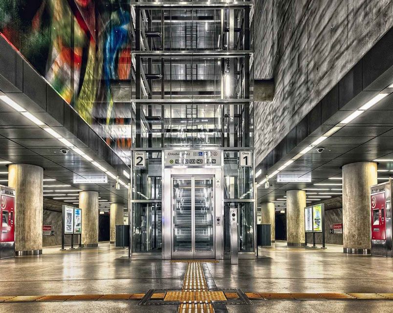 Ascenseur dans une station de métro, aux parois de verre. Des colonnes et des fresques colorées ornent les côtés.
