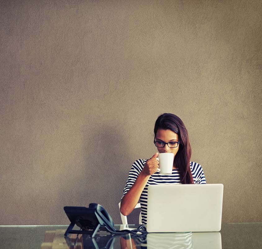 Une femme portant des lunettes boit dans une tasse, travaillant sur un ordinateur portable à un bureau, avec un téléphone à portée de main.