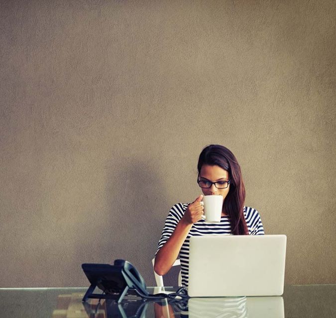 Une femme portant des lunettes boit dans une tasse, travaillant sur un ordinateur portable à un bureau, avec un téléphone à portée de main.
