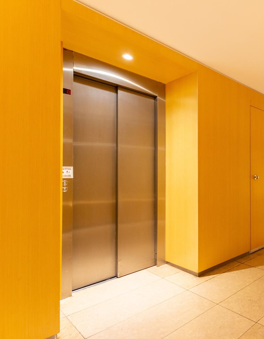 Ascenseur aux portes métalliques fermées dans un couloir aux murs jaunes et au sol carrelé beige.