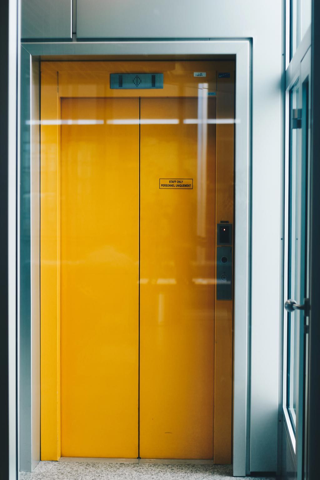 Portes d'ascenseur jaunes à cadre argenté, dans un couloir vitré et blanc.