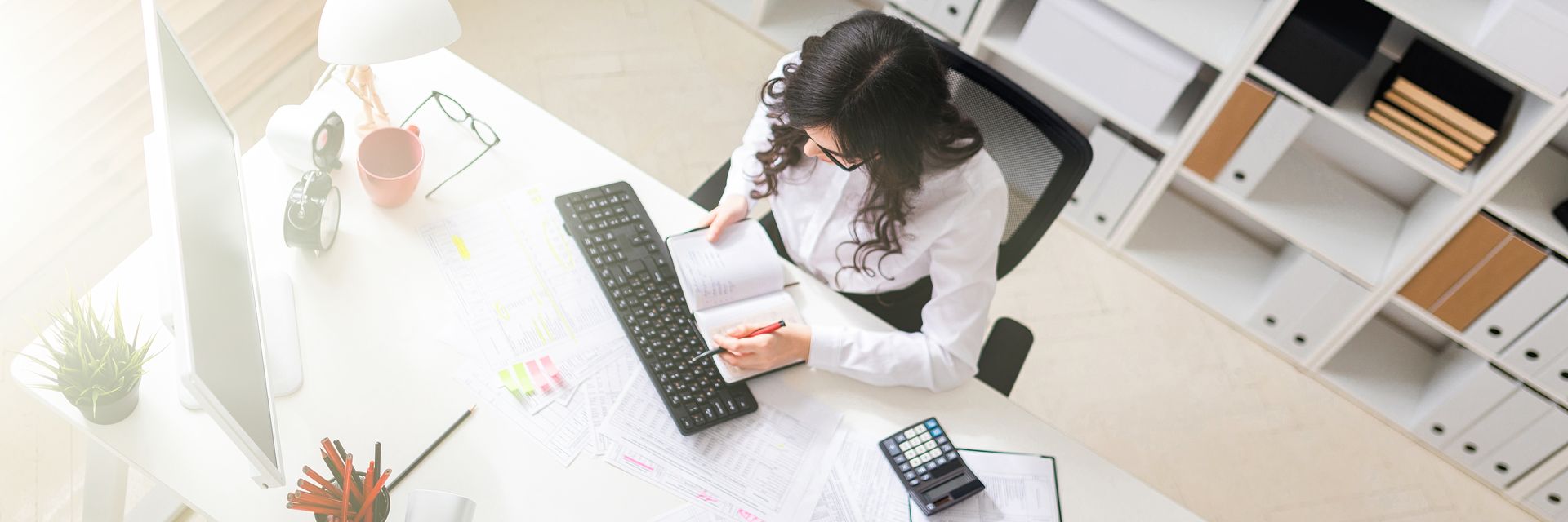 Une personne écrit dans un bureau lumineux, avec un clavier, des papiers et des étagères à proximité.