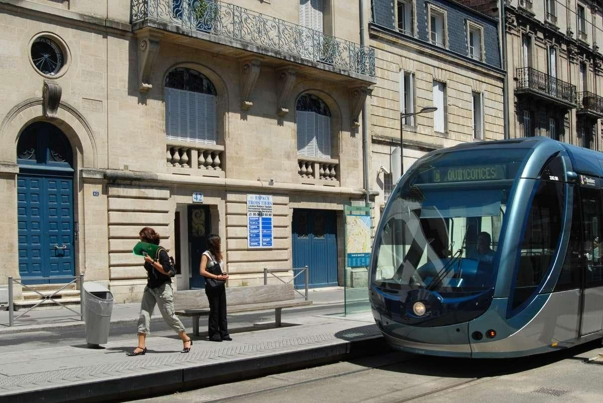 Scène de rue avec un tramway bleu arrêté devant un bâtiment en pierre et deux piétons sur le trottoir.