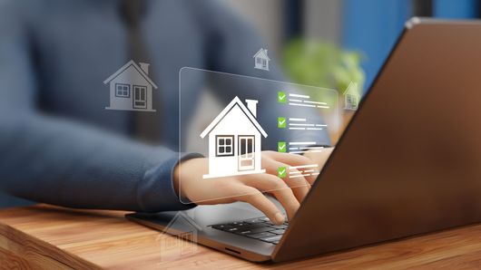 Une personne tape sur un ordinateur portable tandis qu'une superposition numérique d'icônes de maison et d'une liste de contrôle apparaît dans l'air.