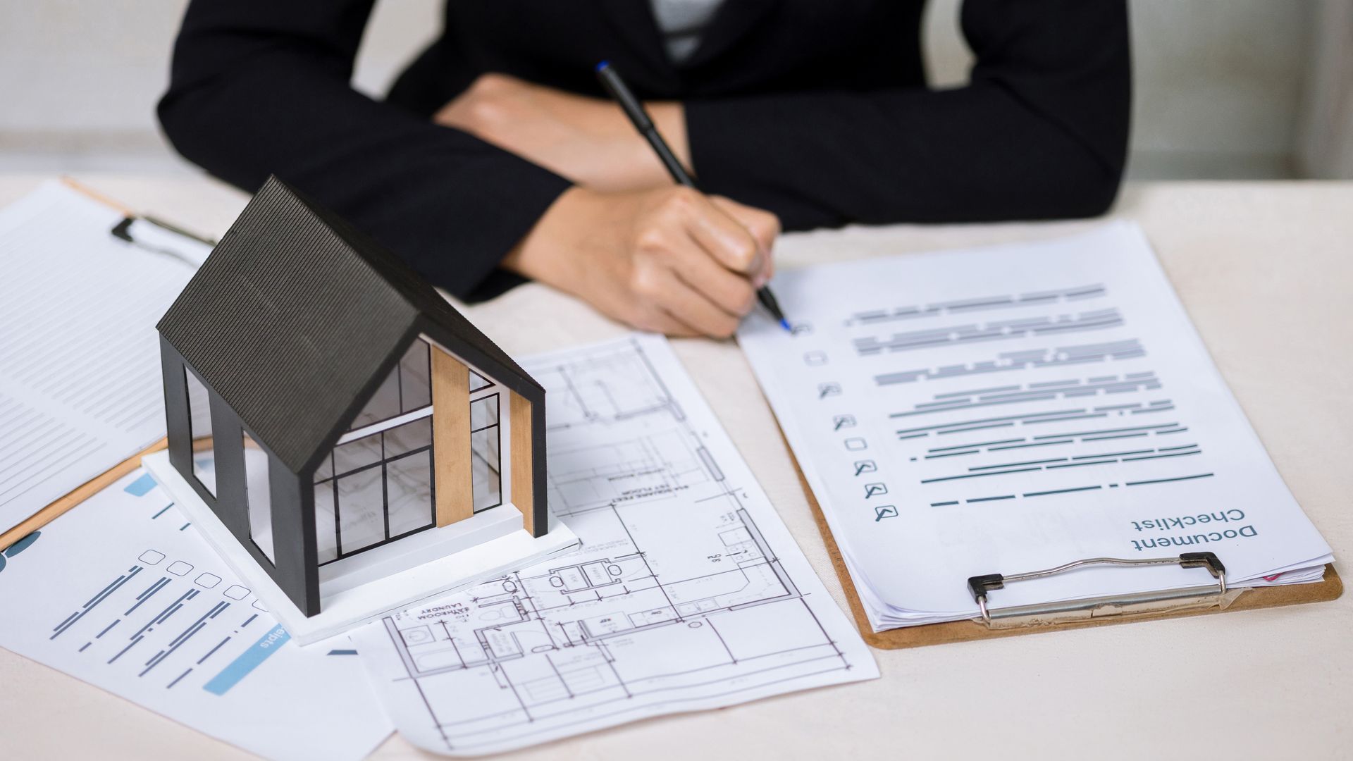 Une personne en costume examine des documents à côté d'une maquette de maison et de plans d'étage posés sur un bureau.