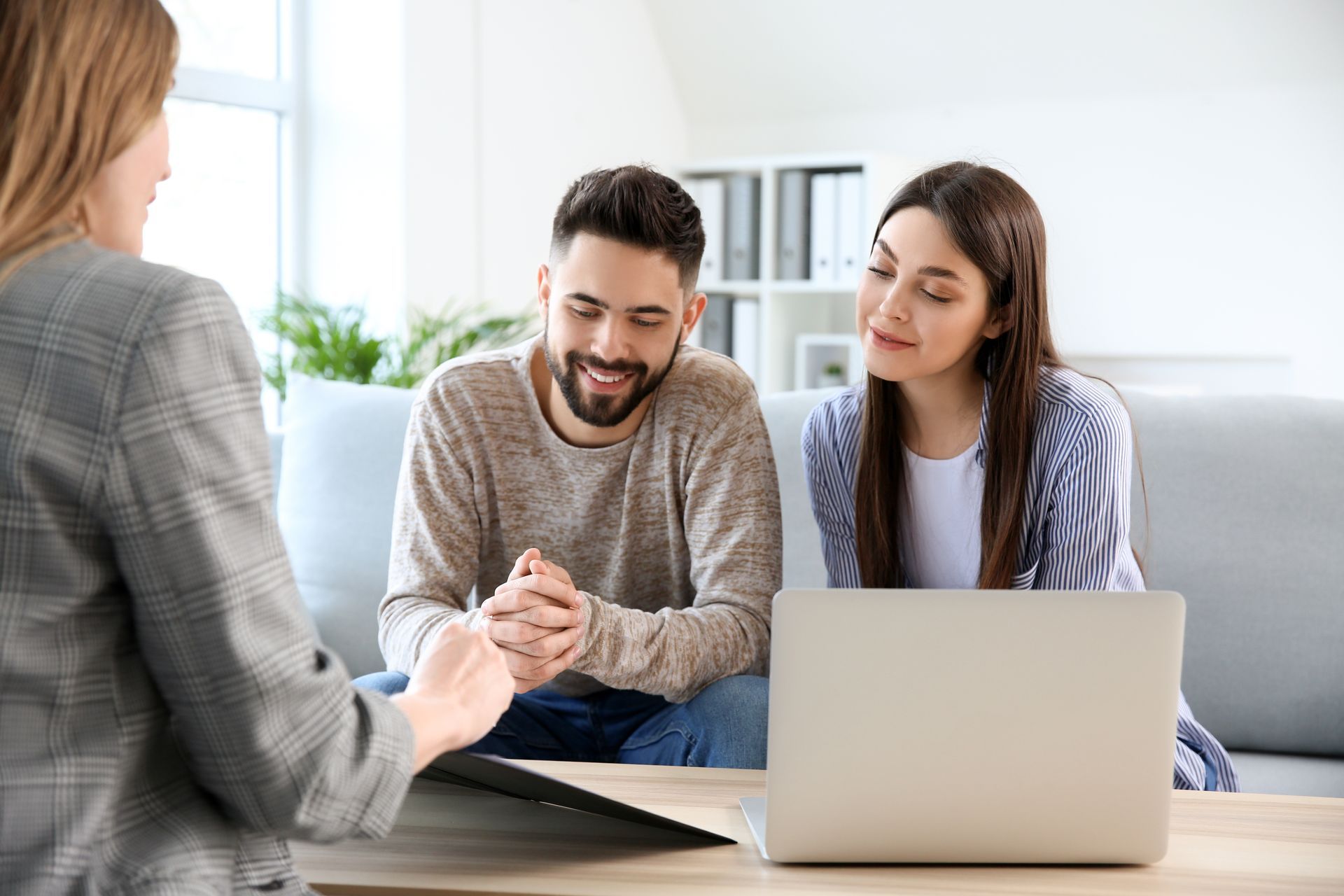 Dans un bureau lumineux, une professionnelle est assise en face d'un couple et ils examinent ensemble des documents devant un ordinateur portable ouvert.