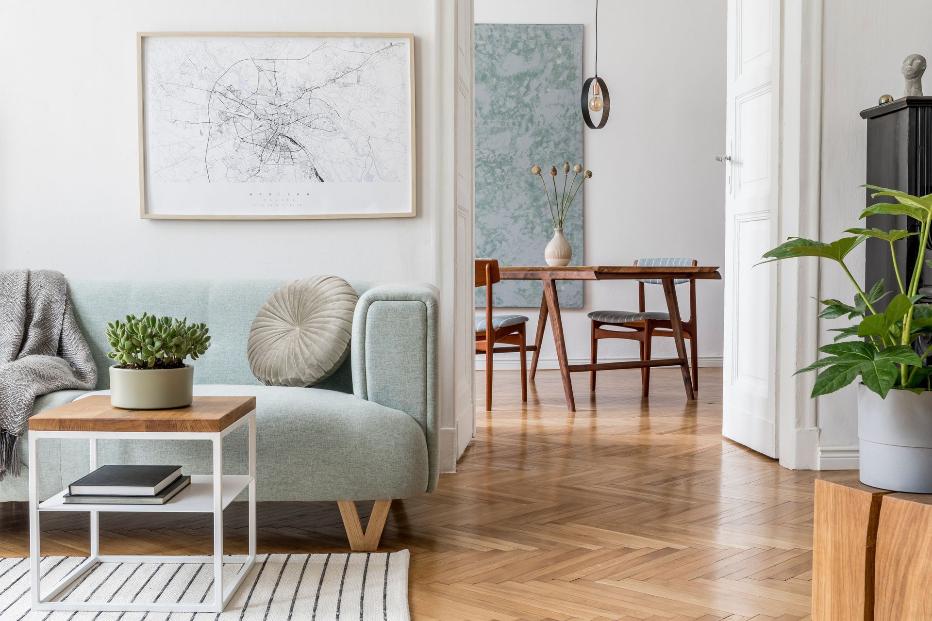 Un canapé vert menthe, une table d'appoint avec une plante et une vue sur une salle à manger avec une table en bois et un parquet à chevrons.