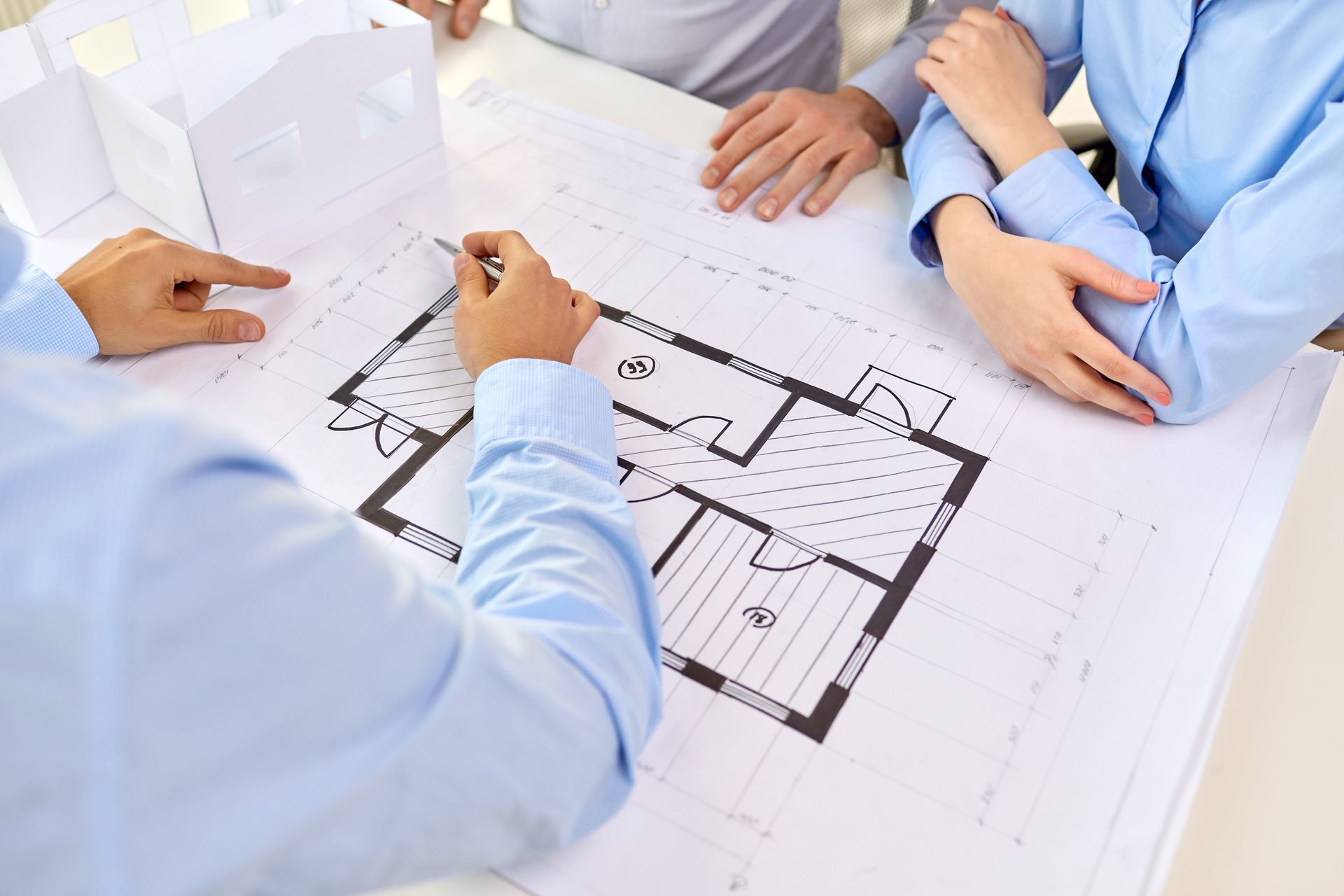 Trois personnes vêtues de chemises bleu clair examinent un plan d'étage résidentiel et une maquette architecturale blanche posés sur une table blanche.