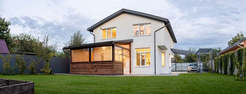 Maison blanche à deux étages avec une terrasse en bois, de grandes fenêtres et une pelouse verte sous un ciel nuageux.