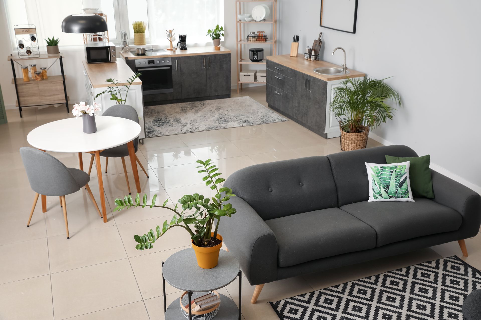 Vue en plongée d'un appartement moderne avec un canapé gris, une table ronde blanche et une cuisine aux tons sombres, sans cloison.