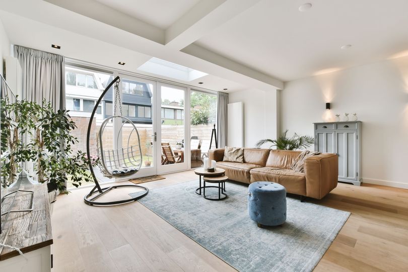 Un salon moderne avec un canapé beige, un pouf bleu, un fauteuil suspendu et de grandes portes vitrées donnant sur une terrasse extérieure.