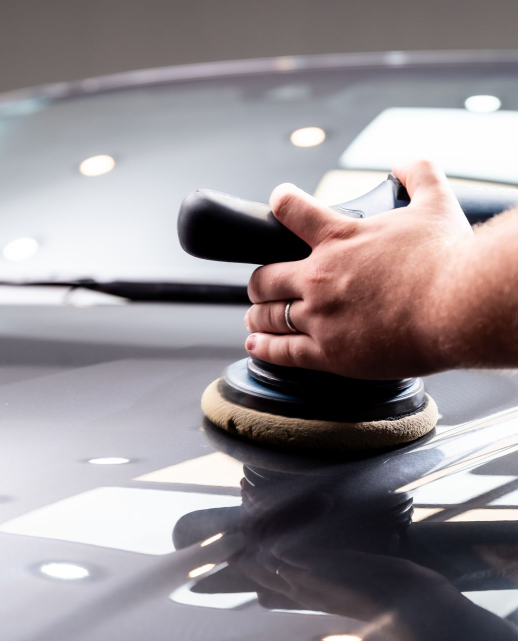 Une personne polit une voiture avec une polisseuse électrique dans un environnement bien éclairé ; on aperçoit sa main qui tient l'outil.