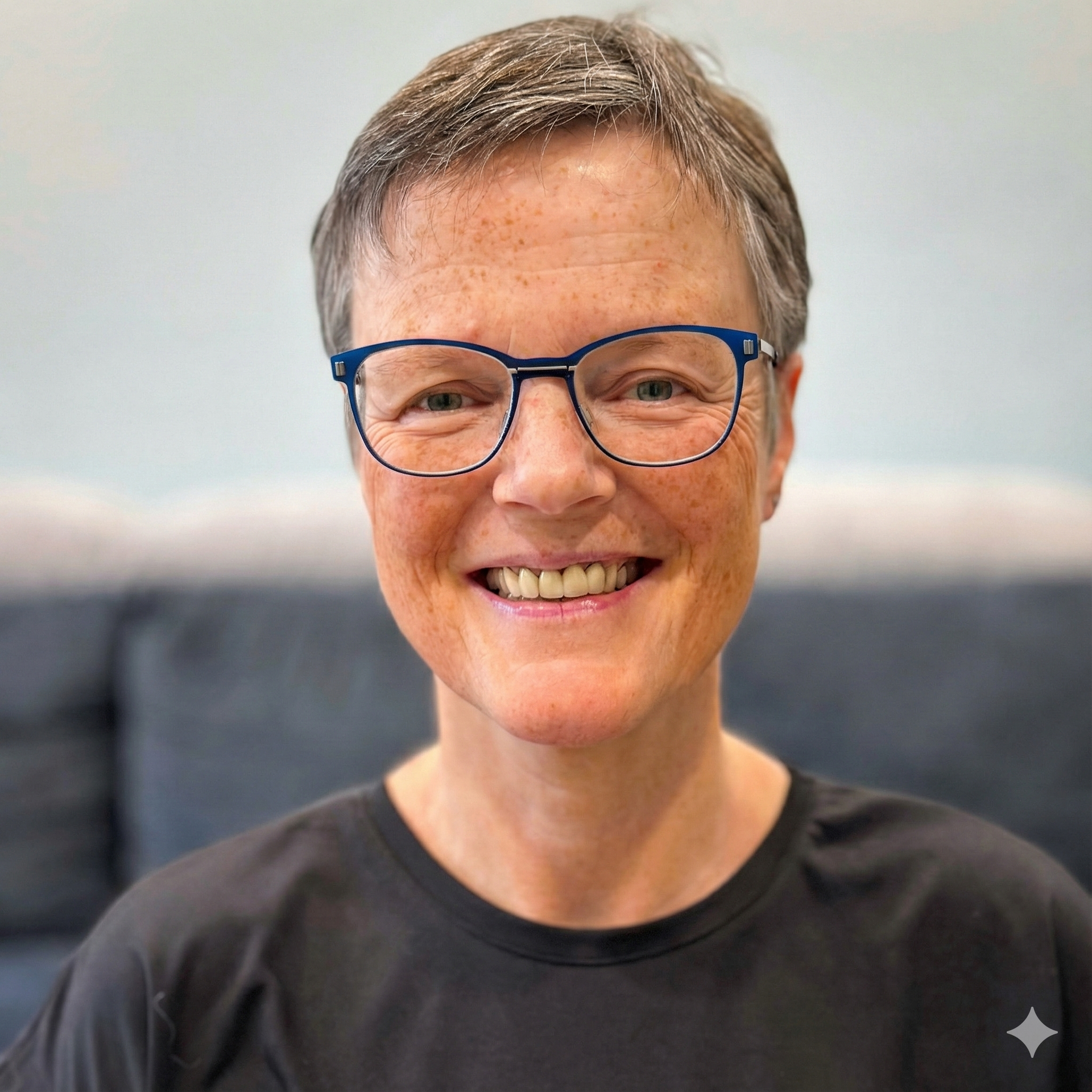 Eine Person mit kurzen grauen Haaren und blau umrandeter Brille lächelt in die Kamera; sie trägt ein schwarzes Hemd.