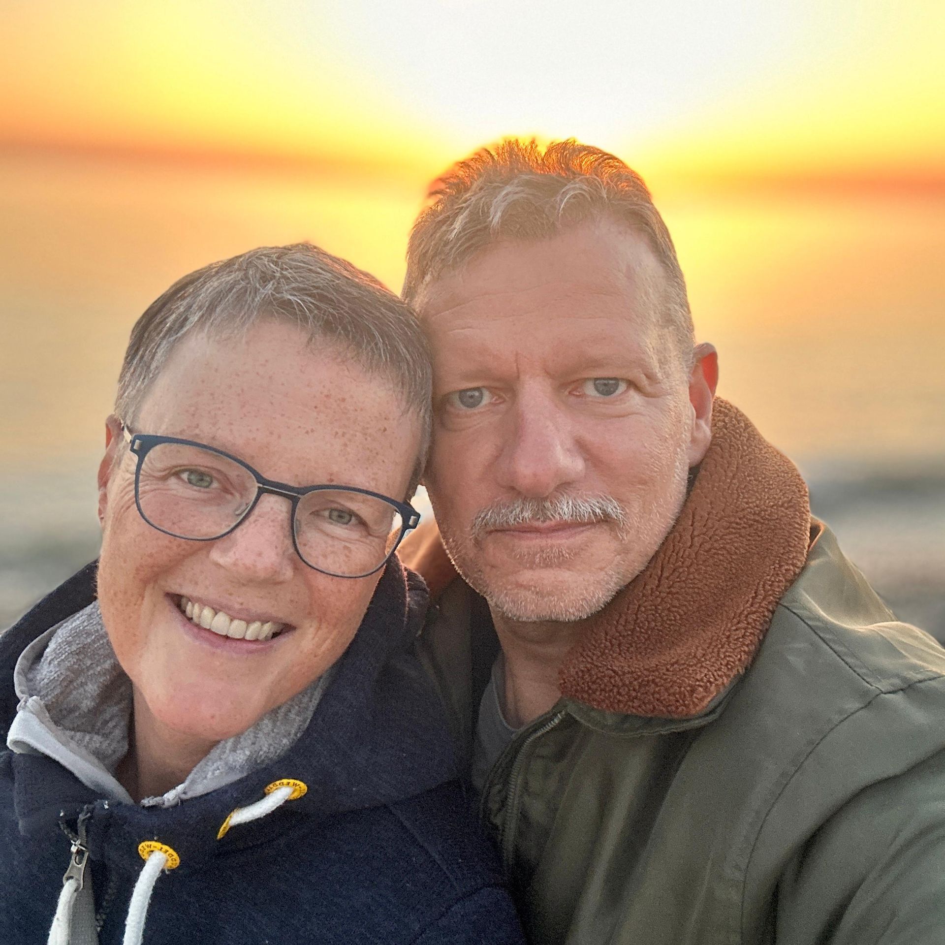 Ein Paar lächelt vor einem Sonnenuntergang über dem Wasser. Er trägt eine Brille und einen Kapuzenpullover, der andere eine Jacke mit Pelzkragen.