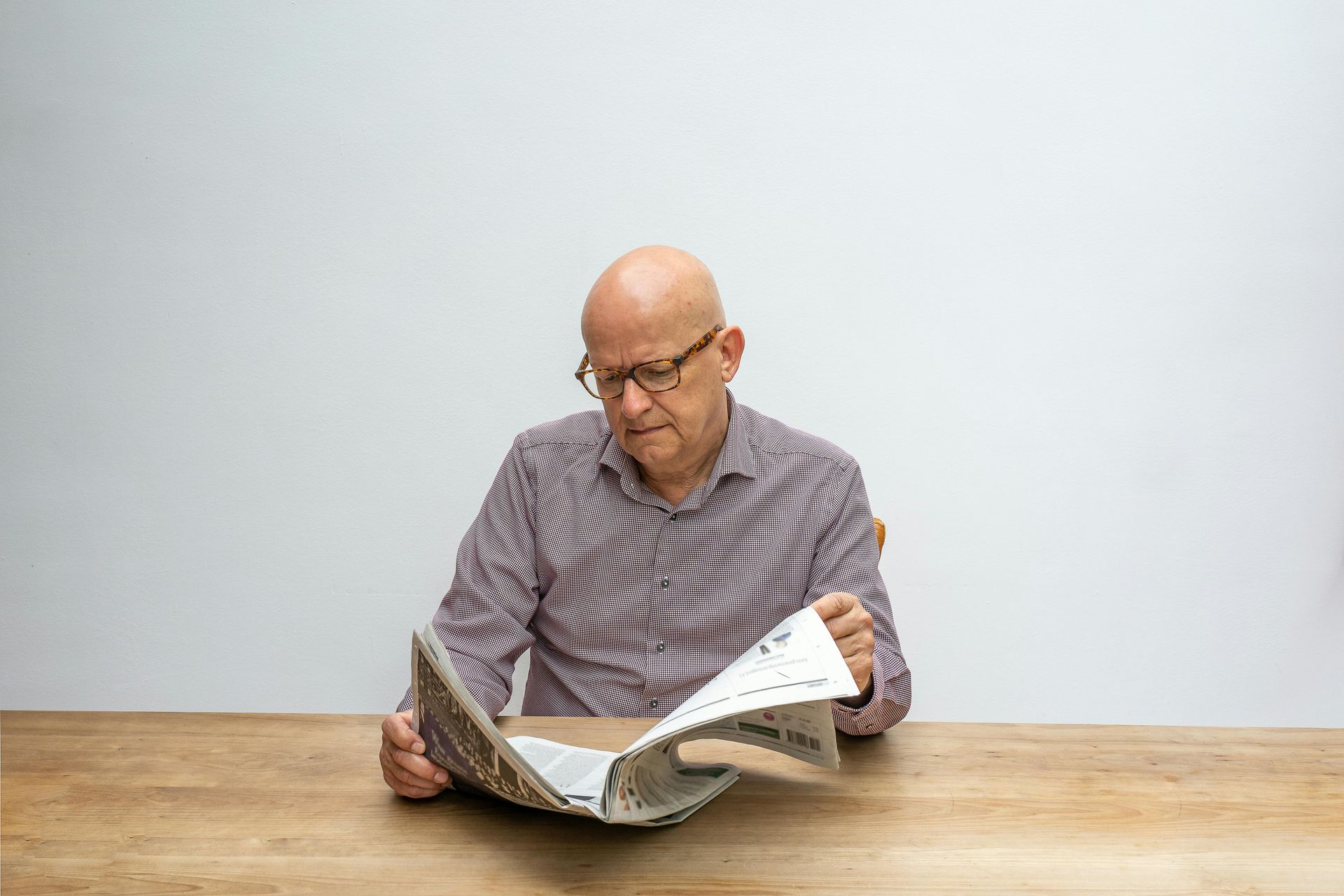 Glatzköpfiger Mann mit Brille liest an einem Holztisch Zeitung.