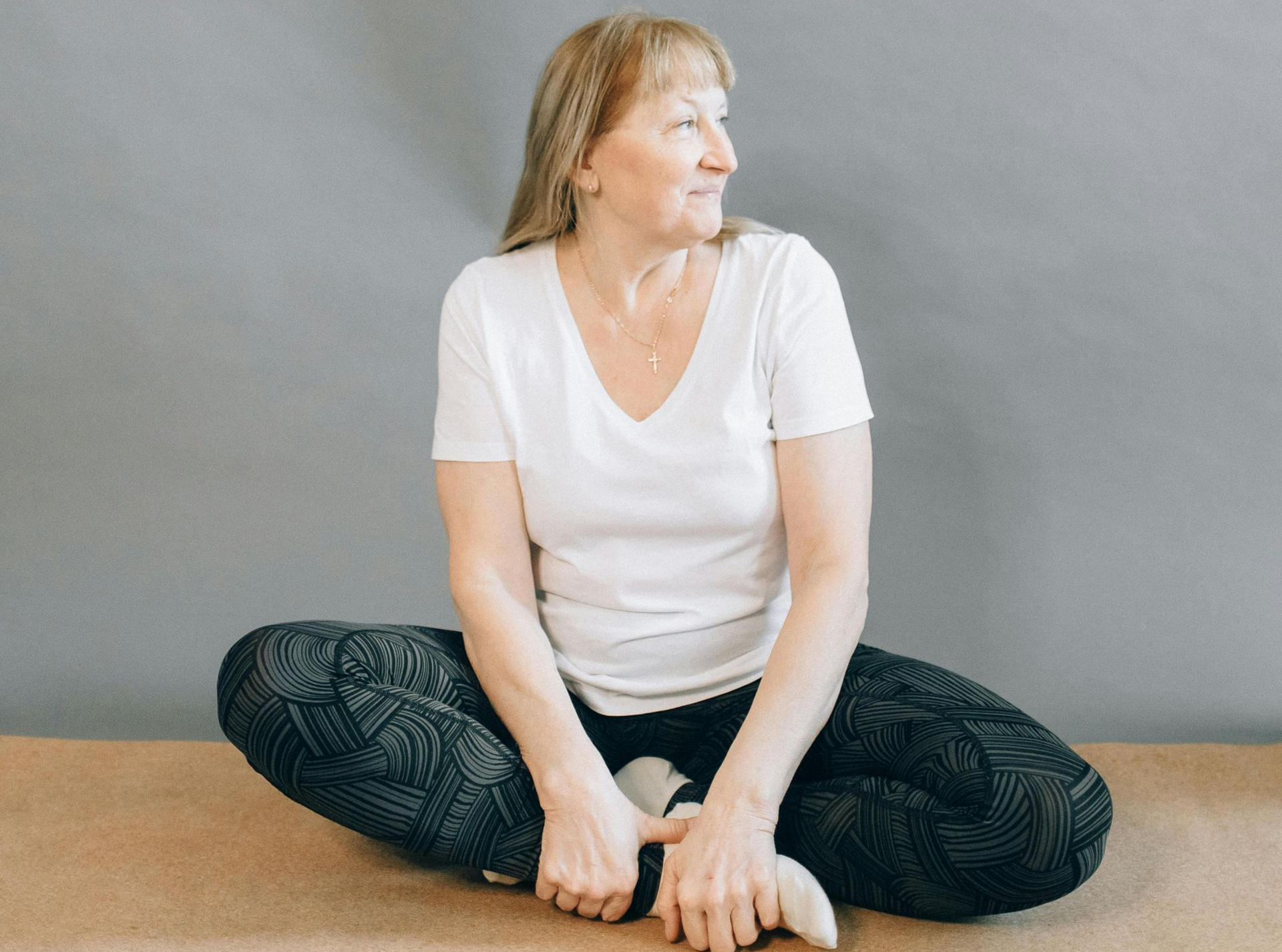 Woman seated cross-legged, looking to the side. Wearing white t-shirt, patterned leggings, on a tan surface. Gray backdrop.