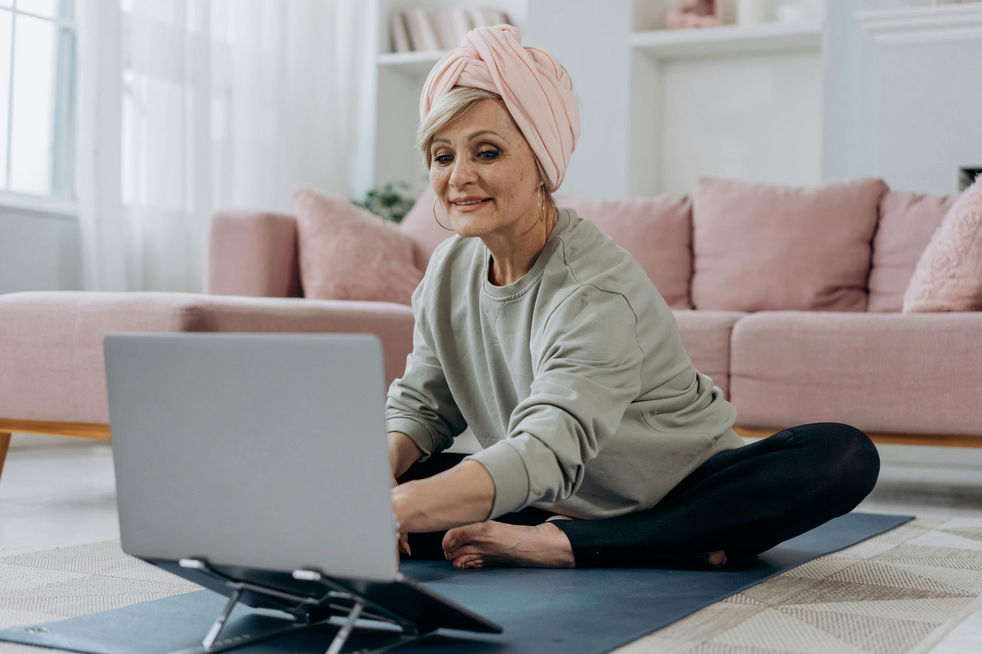 Eine Frau mit rosa Turban sitzt im Wohnzimmer im Schneidersitz auf einer Yogamatte, schaut auf ihren Laptop.