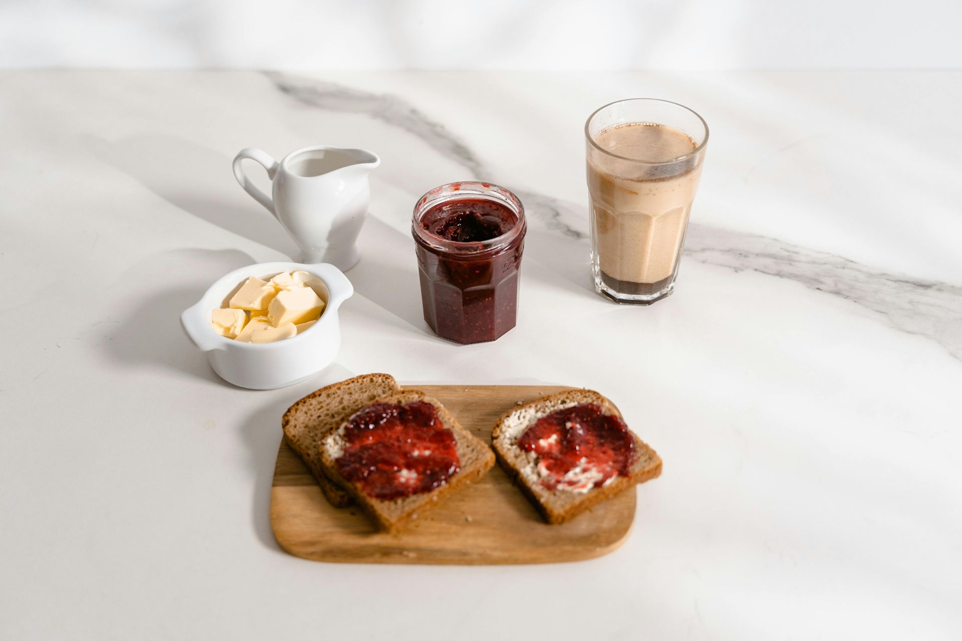 Frühstück: Toast mit Marmelade und Butter, Kaffee, Marmeladenglas, Milchkännchen und Butterdose auf einer Marmorplatte.