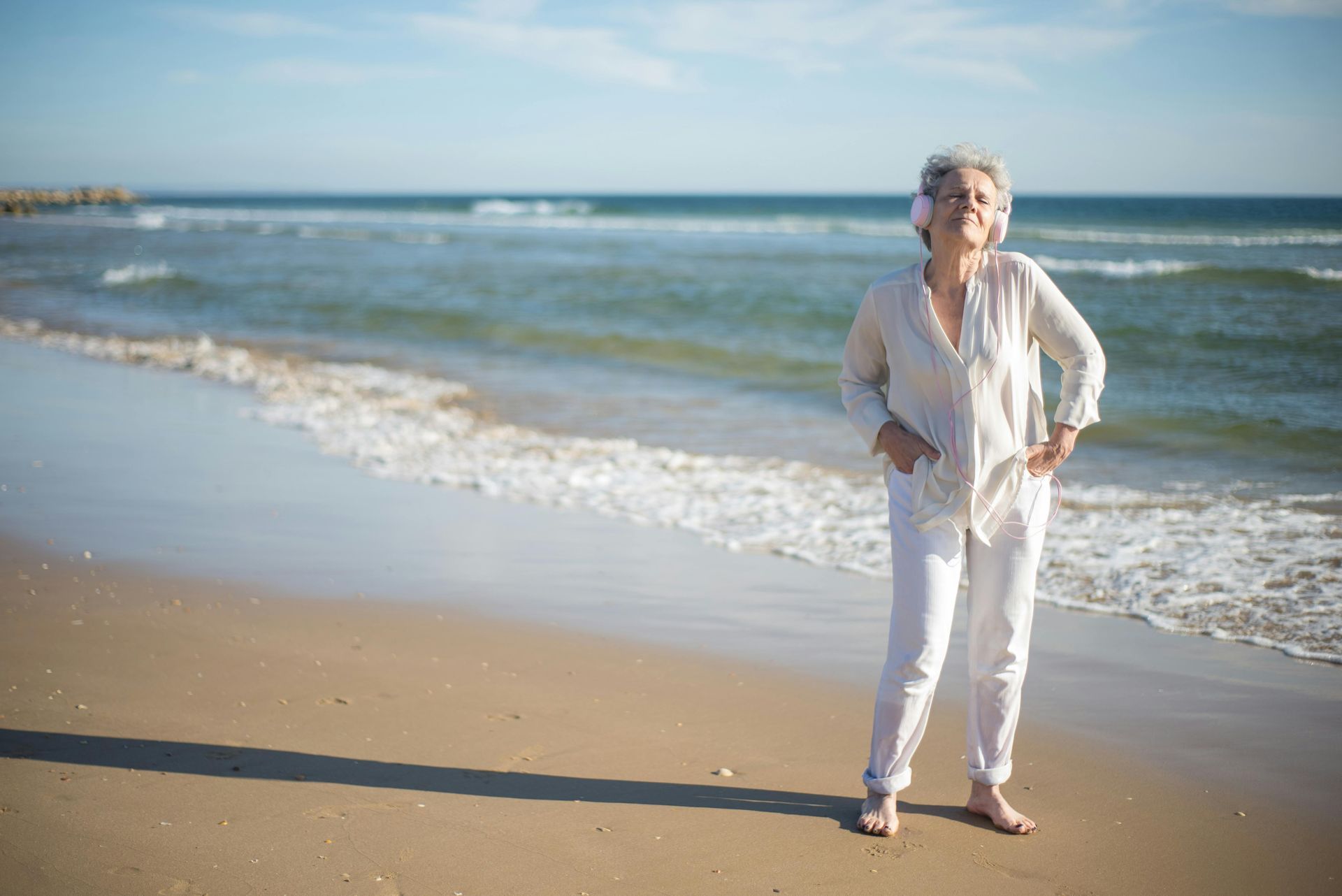 Eine Frau in weißem Outfit steht am Strand, die Hände in den Taschen, und blickt aufs Meer.
