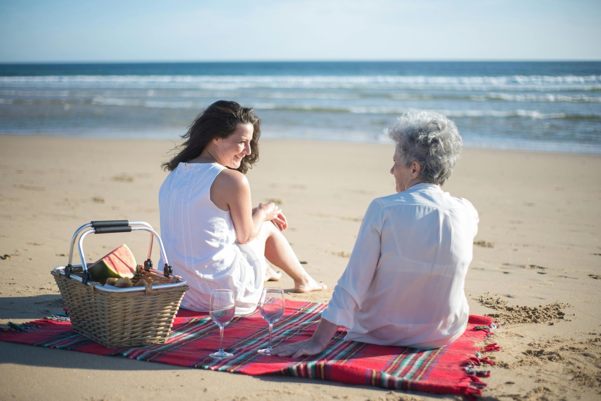 Zwei Personen sitzen auf einer Stranddecke und schauen einander an; Picknickkorb, im Hintergrund das Meer.