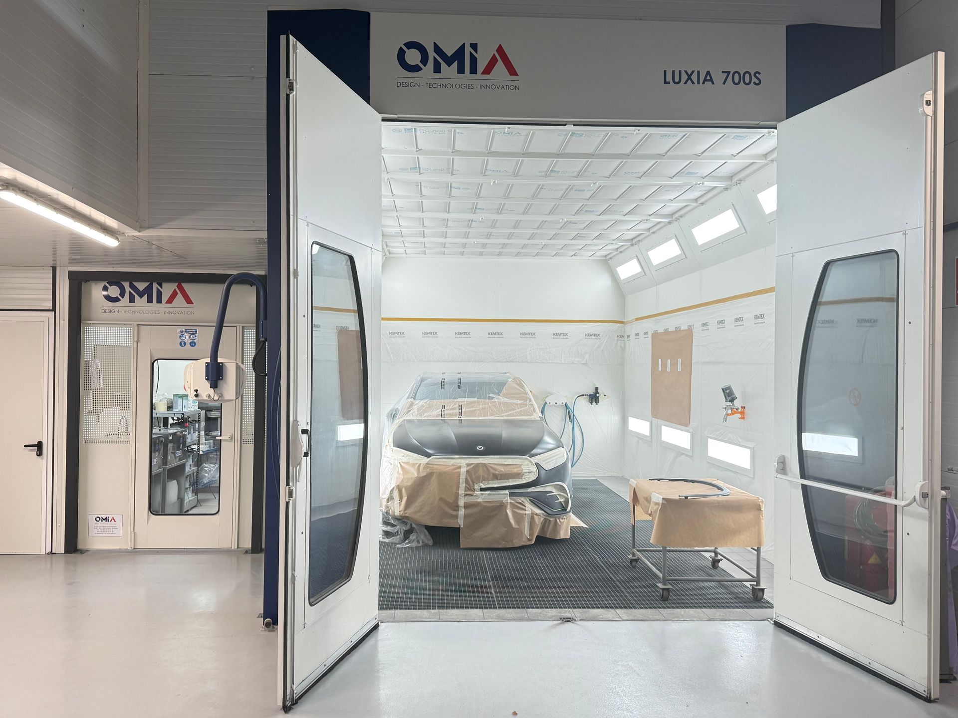 Cabine de peinture avec une voiture à l'intérieur, portes ouvertes. L'intérieur est blanc avec des éclairages au plafond et un sol noir.