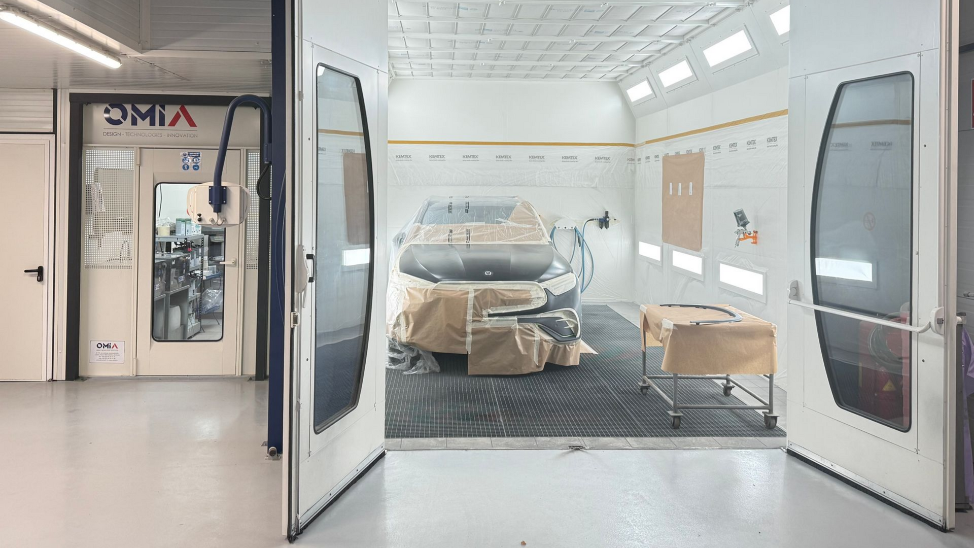 Cabine de peinture avec une voiture à l'intérieur, portes ouvertes. L'intérieur est blanc avec des éclairages au plafond et un sol noir.