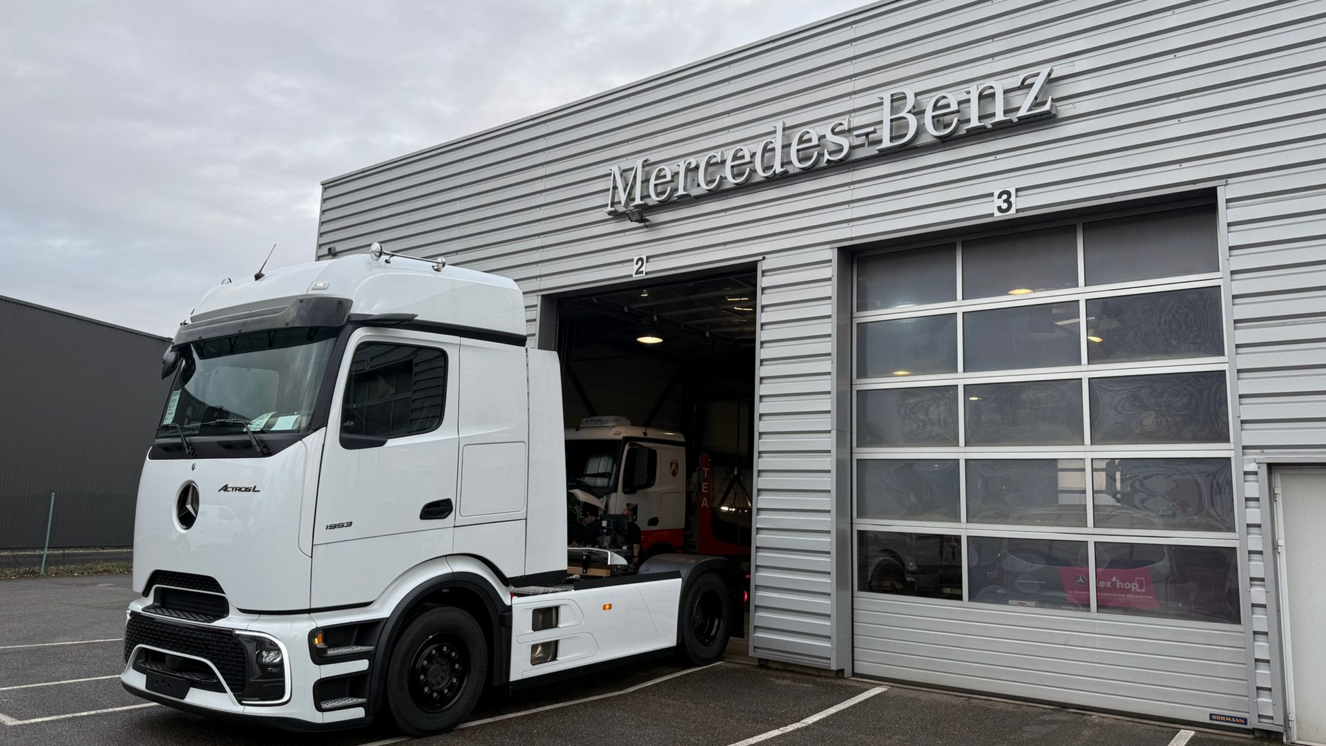 Camion blanc stationné sur le parking d'un centre de service Mercedes-Benz, porte de garage entrouverte.