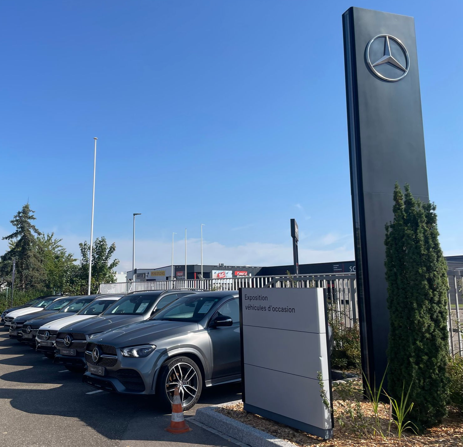 Extérieur du garage Mercedes-Benz avec des voitures garées en rang, devant une grande enseigne totem arborant le logo Mercedes.