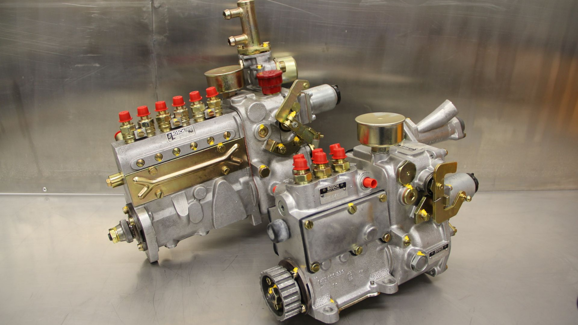 Deux pompes à carburant argentées avec raccords en laiton et capuchons rouges sur une surface en acier inoxydable.