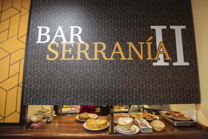 Letrero del Bar Serranía sobre un mostrador con pasteles y bandejas de comida exhibidas debajo.