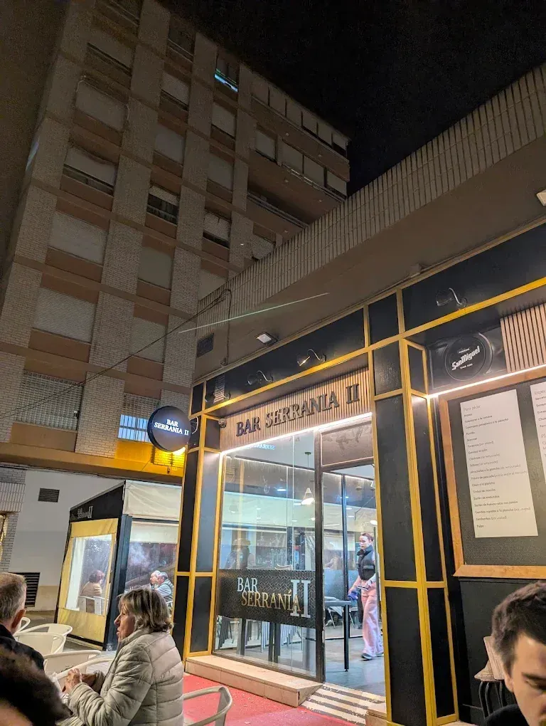 Exterior nocturno de la entrada de un edificio con fachada de cristal, con un letrero iluminado y gente de pie cerca.