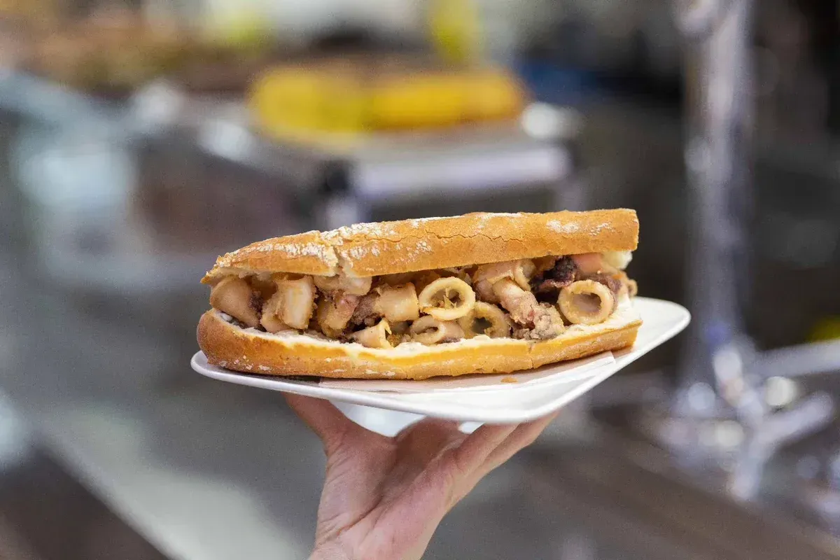 Un sándwich con calamares fritos en un plato blanco, sostenido en la mano en un puesto de mercado.