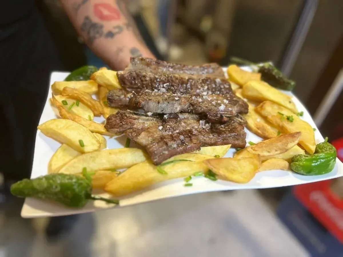 Plato de carne a la parrilla y papas fritas adornado con pimientos verdes, sostenido por una persona al fondo.