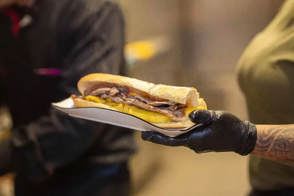 Una mano enguantada sostiene un sándwich de cheesesteak en una bandeja blanca en un entorno de cocina.